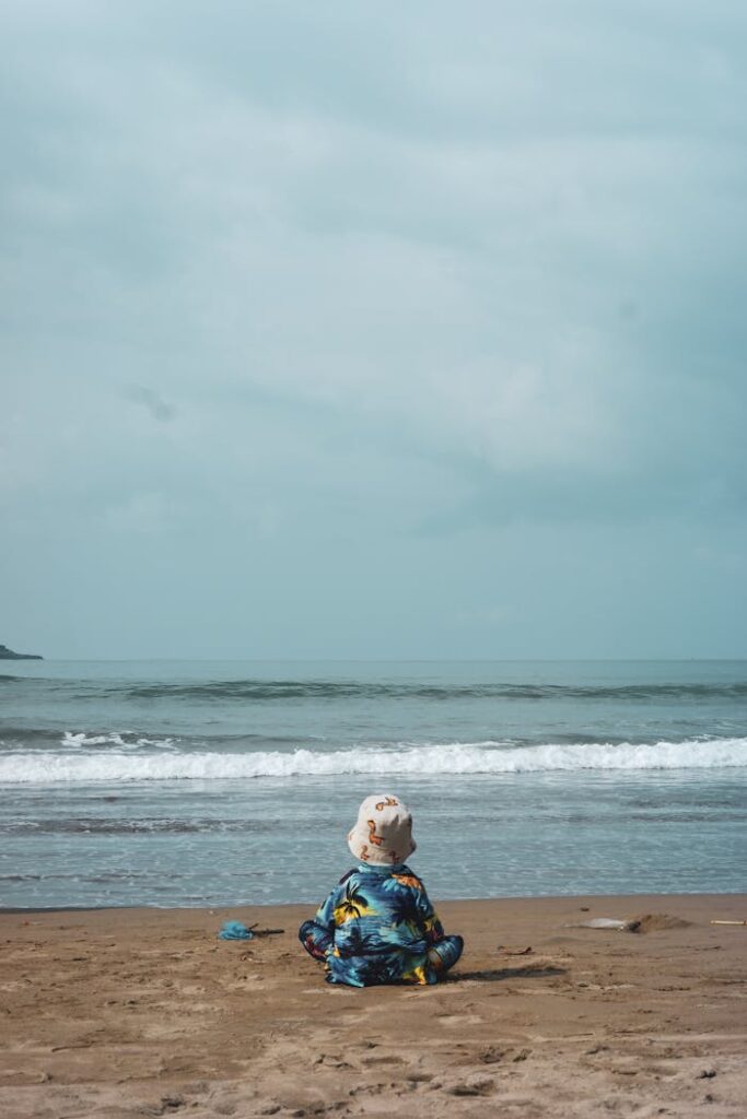 A child with a hat sits on a peaceful beach, gazing at the ocean waves.