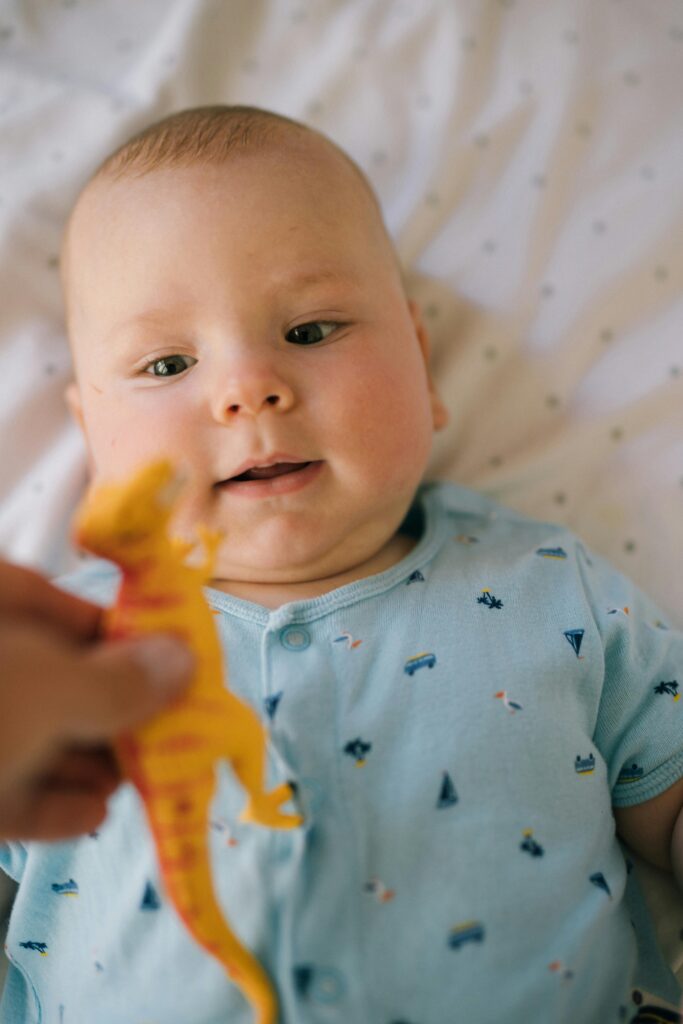 Cute baby in a blue outfit playing with a dinosaur toy on a star-patterned bed sheet.